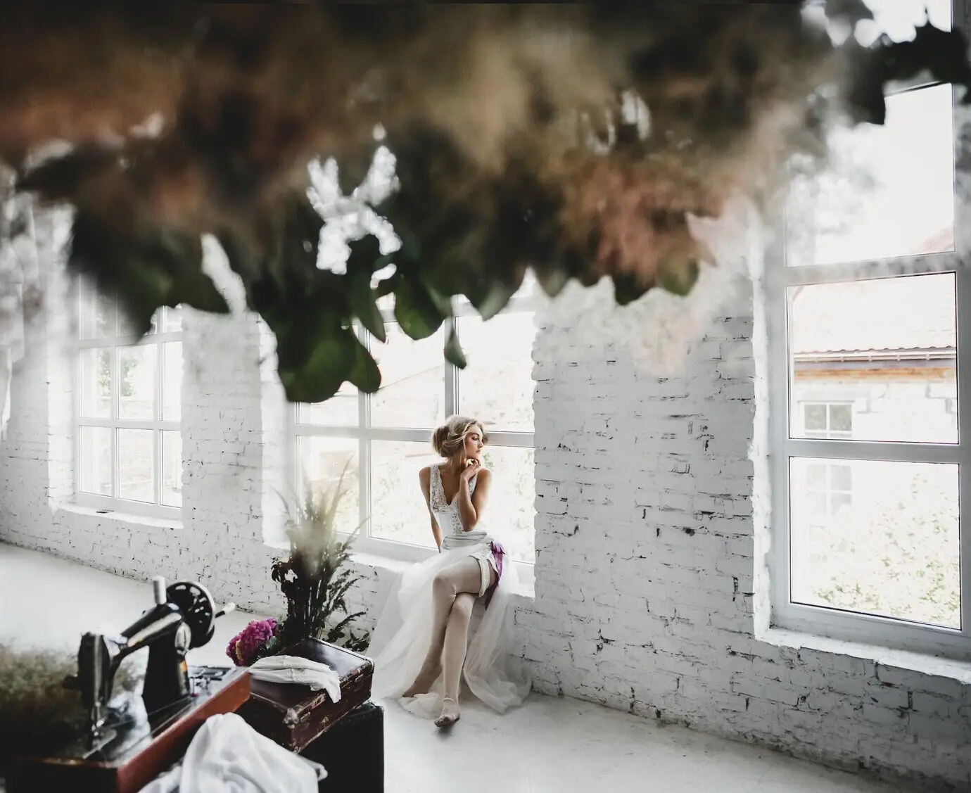 Женщина в белой одежде сидит на подоконнике в комнате с цветами и швейной машиной.